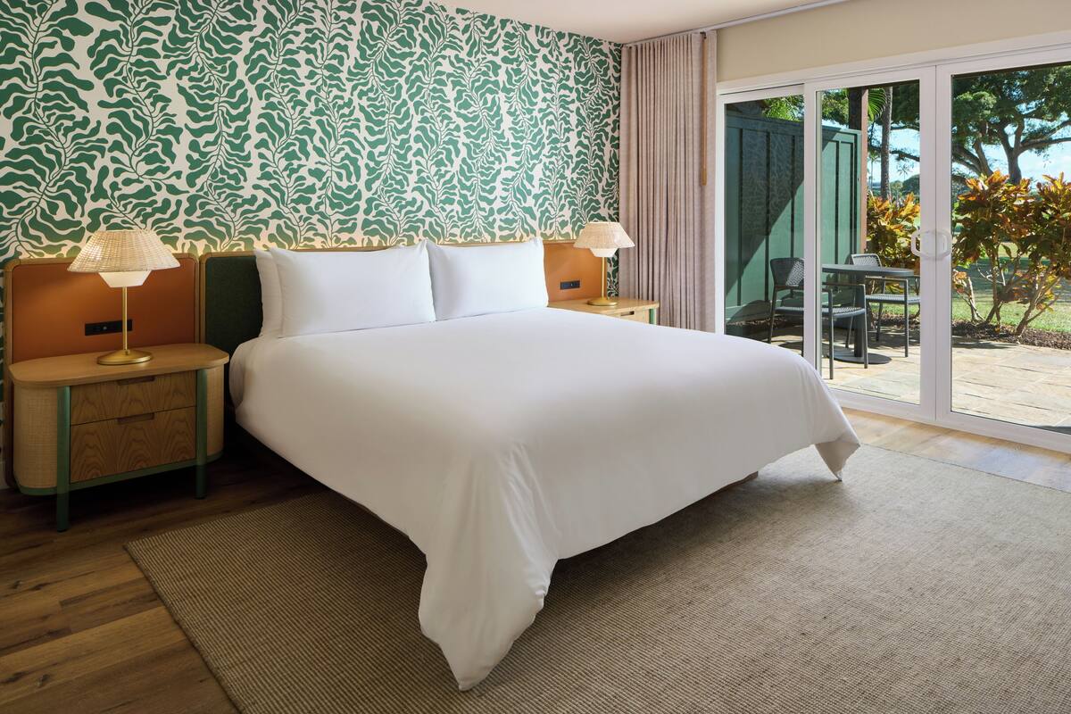 Habitación de hotel con una cama grande, dos mesitas de noche con lámparas, papel tapiz estampado y una puerta corrediza de vidrio que da a un espacio exterior.