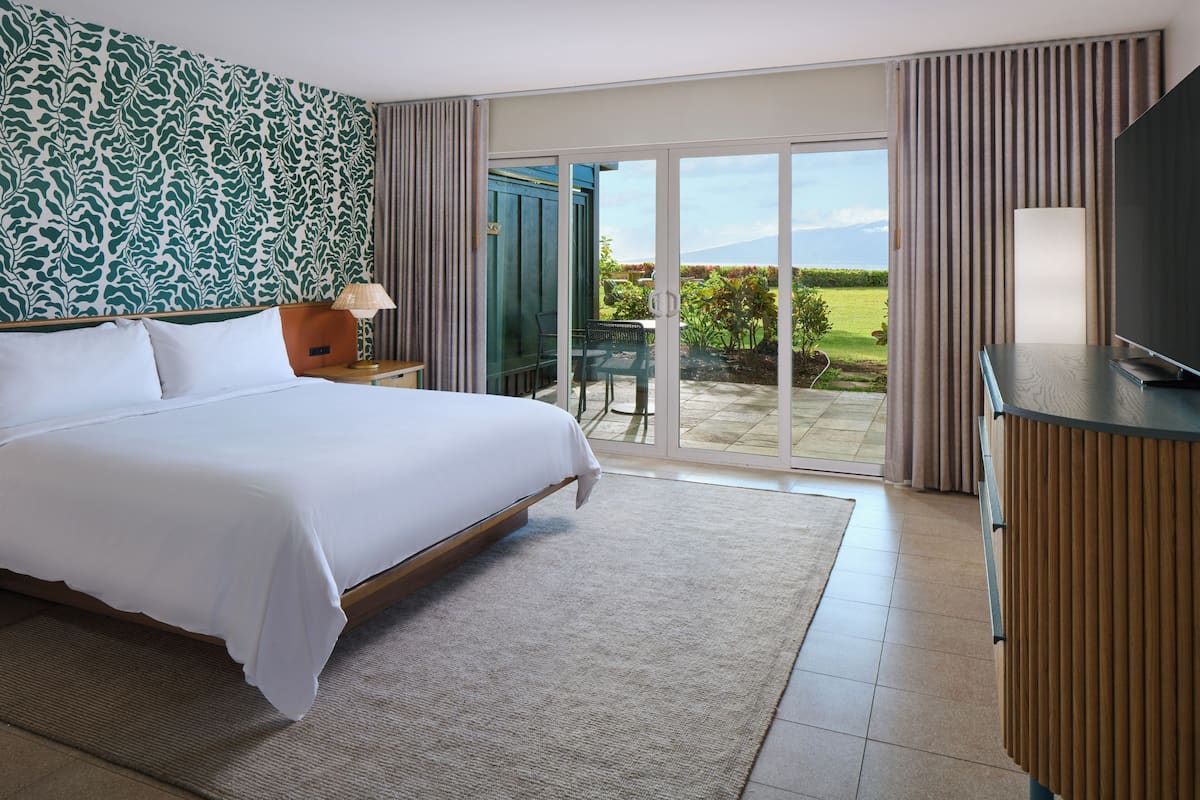 Habitación de hotel con una cama grande, televisión, escritorio y vistas a un jardín por una puerta corrediza de vidrio.