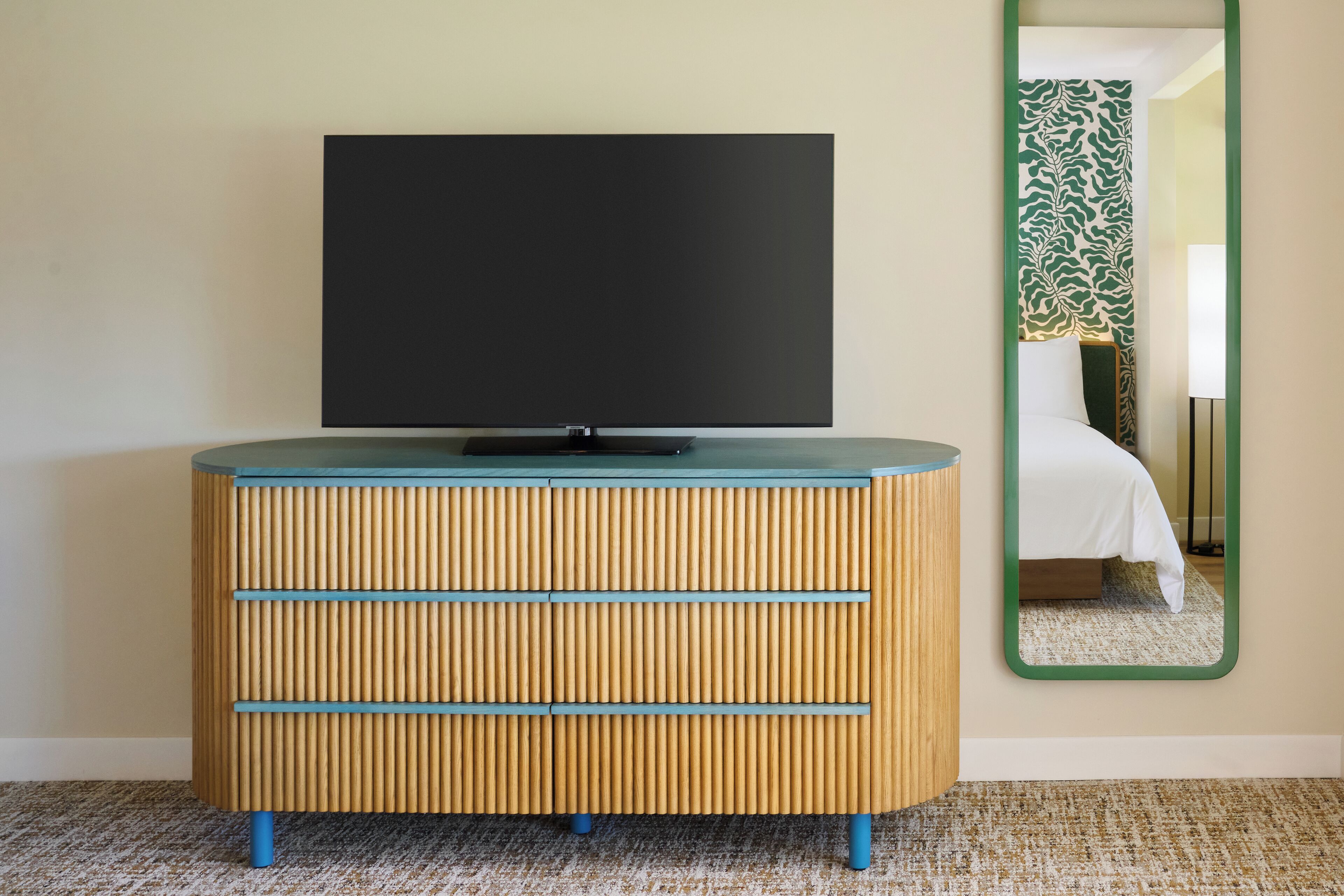 A wooden cabinet with drawers and a flat-screen TV on top, a mirror on the wall, and a bed with a patterned headboard.