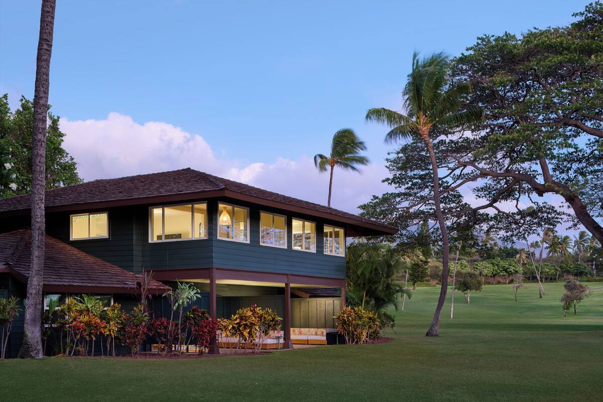 Una casa de dos pisos con porche cubierto, rodeada de césped cuidado y vegetación tropical.