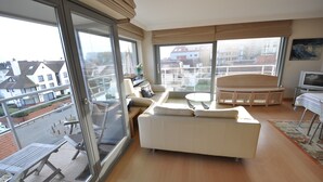 A modern living room with a sofa, a small table, and a TV. Large windows offer a view of the city. - Apartment in Belgium With Side Sea View (Knokke-Heist)