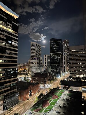 Paisaje urbano nocturno con edificios modernos y altos, una intersección bien iluminada y una zona ajardinada.
