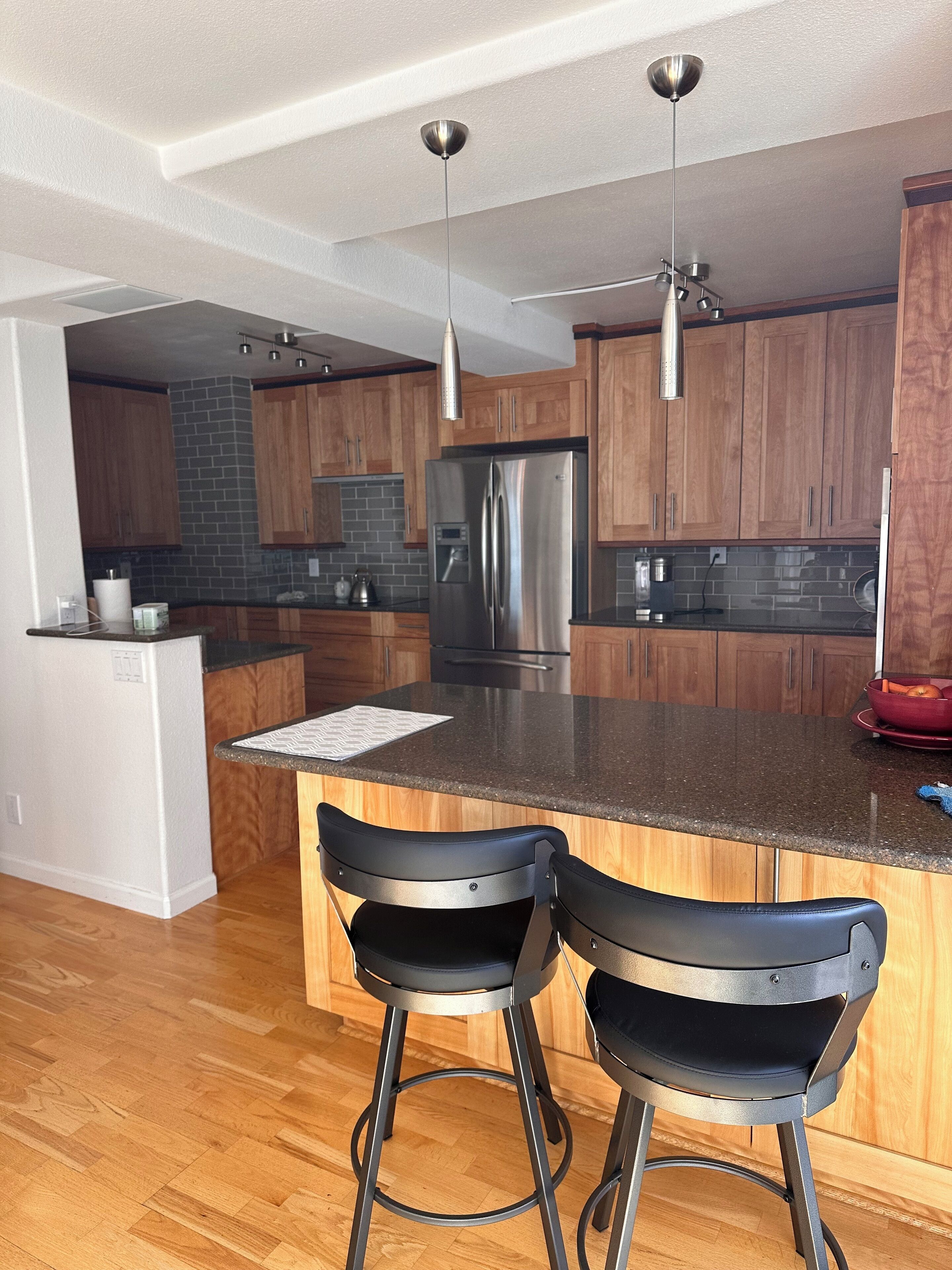 A modern kitchen with wooden cabinets, a stainless steel refrigerator, and two black bar stools.