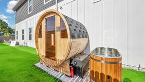 A wooden sauna structure with a large door, a metal container, and a fire extinguisher on a grassy lawn. - Luxury Beach Home Sauna Cold Plunge E-bikes (Jacksonville Beach)