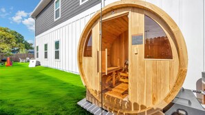 A wooden sauna structure with a bench inside, attached to a house with white siding and a green lawn. - Luxury Beach Home Sauna Cold Plunge E-bikes (Jacksonville Beach)