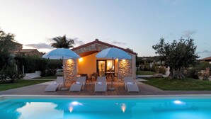 A poolside area with lounge chairs and umbrellas, a stone wall, and a building with a tiled roof. - Casa Moros 6, Emma Villas (Castiadas)