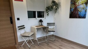 A room with a wooden floor, a desk with two chairs, a window with blinds, a door, and a wall-mounted light fixture. - Apartamento moderno cerca del aeropuerto Tenerife Norte y La Laguna. (CN)