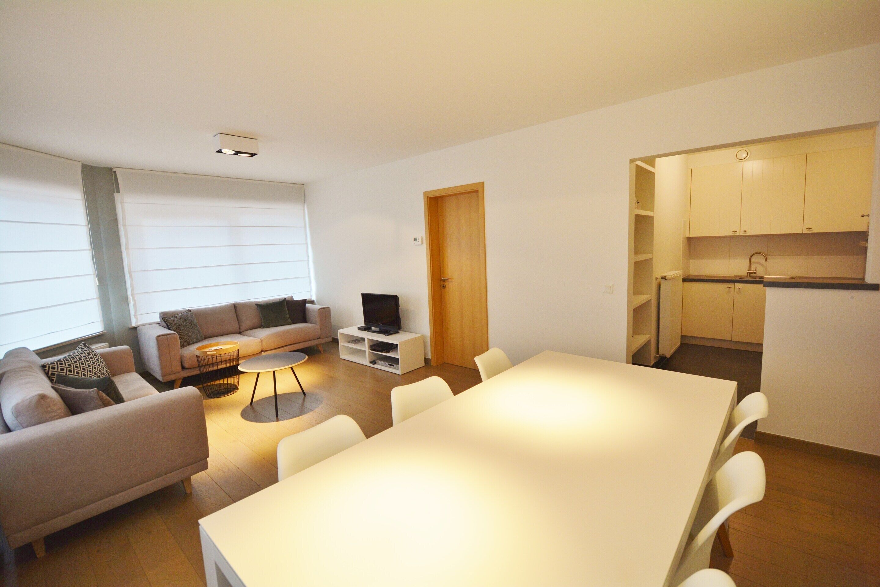 Una sala moderna con sofá, mesa de centro y televisor, un comedor con mesa y sillas, y una cocina con armarios y pileta.