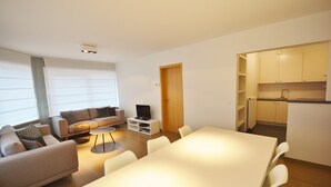 A modern living room with a sofa, coffee table, and TV, a dining area with a table and chairs, and a kitchen with cabinets and a sink. - Apartment in Knokke Near the Beach (Knokke-Heist)