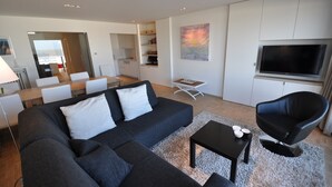 A modern living room with a sofa, coffee table, and a TV. There is a dining area with chairs and a table. The room has white walls and a kitchen area with cabinets and a microwave. - Apartment in Duinbergen by Sailing Club (Knokke-Heist)