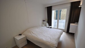 A bedroom with a large bed, a small bedside table, a chair, a radiator, and a window with curtains. - Apartment in Duinbergen by Sailing Club (Knokke-Heist)