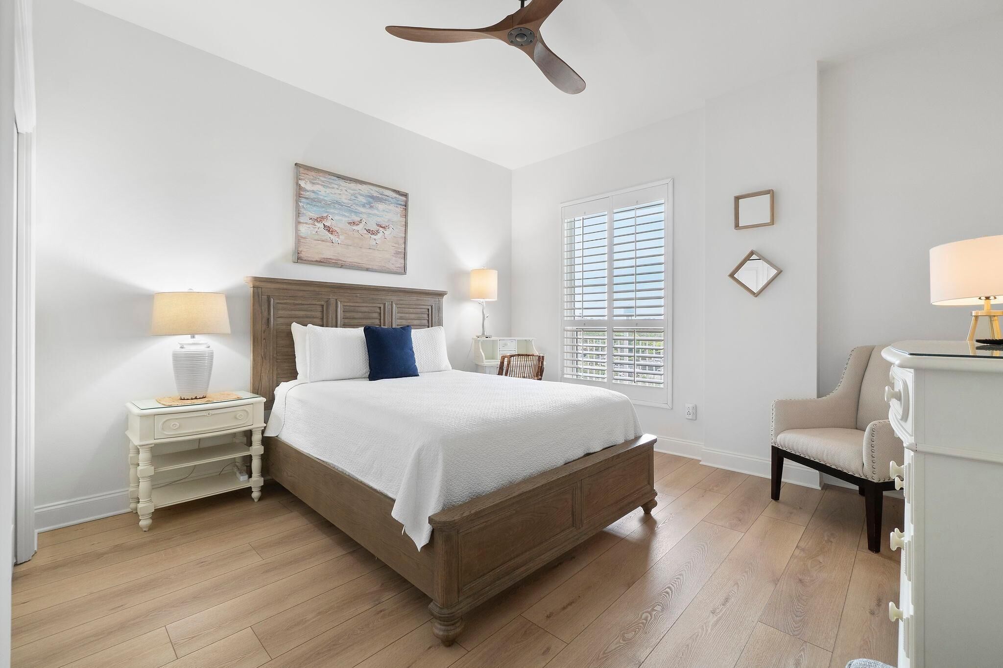 Quarto com cama de madeira, duas mesas de cabeceira com abajures, ventilador de teto, janela com persianas e uma cadeira.