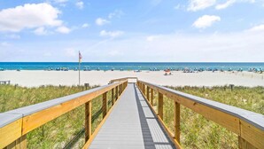 Ein hölzerner Steg führt zu einem Strand mit Sonnenschirmen und Menschen.