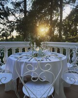 Uma mesa de jantar redonda posta para uma refeição, com toalhas brancas e cadeiras de metal, cercada por um parapeito branco, com árvores e luz solar ao fundo.