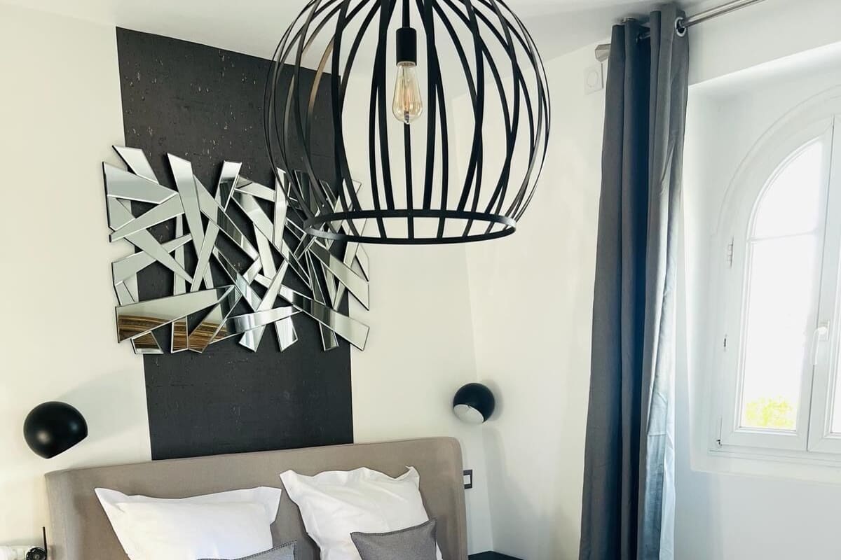 Ein modernes Schlafzimmer mit einem großen Bett, einem geometrischen Wandbild, einer Pendelleuchte und einem Fenster mit Vorhängen.