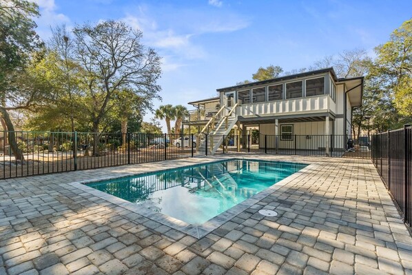 A swimming pool with a paved deck, surrounded by a fence, and a two-story building with a covered patio area in the background.