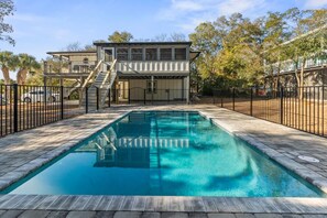 A rectangular swimming pool with a wooden deck surrounding it, a two-story building with a balcony in the background, and a black metal fence enclosing the area.