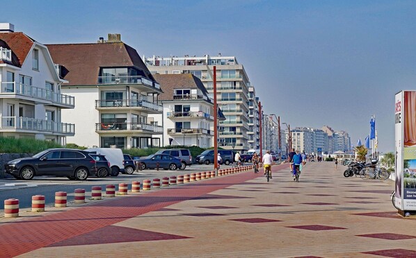 A coastal promenade with modern buildings, a bike lane, and parked cars.