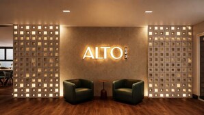 Een moderne lobby met een bord aan de muur waarop 'ALTO N.AHR' staat, twee groene fauteuils, een kleine ronde tafel en een houten vloer.