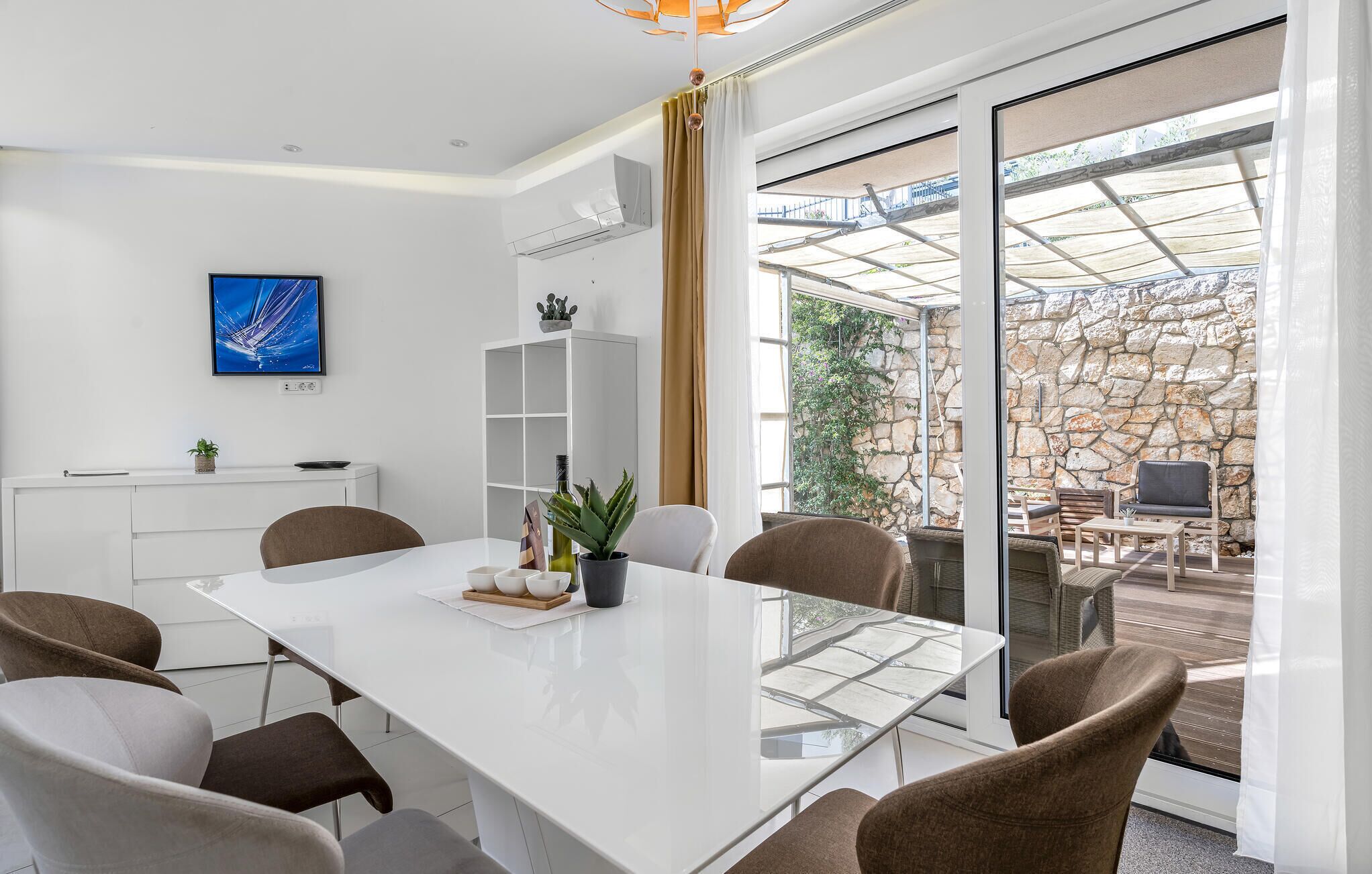 A modern dining area with a white table, brown chairs, and a glass partition. There is a potted plant on the table and a view of an outdoor patio with stone walls and furniture.