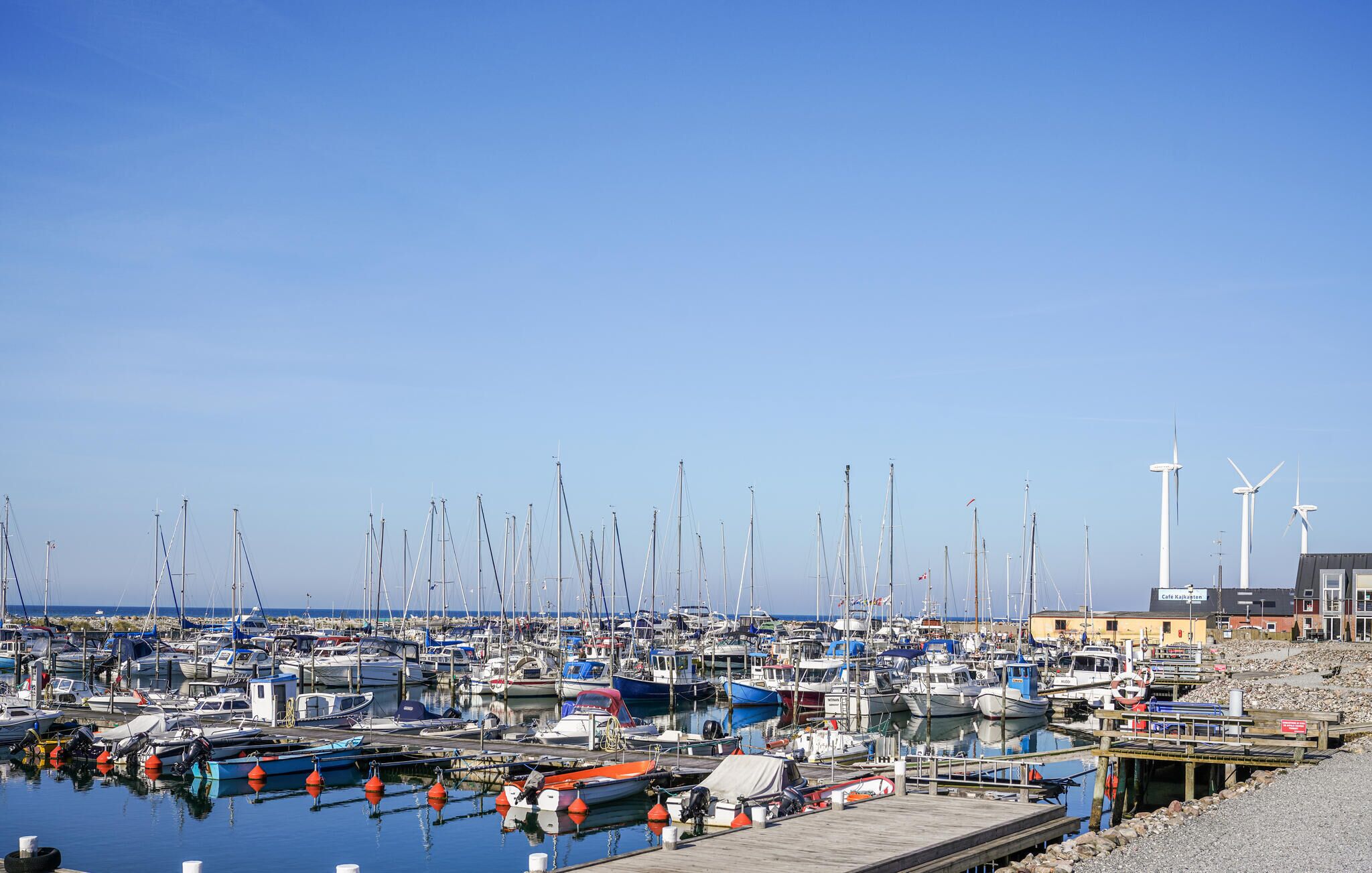 A marina with numerous boats docked, a wooden pier, and wind turbines in the background.
