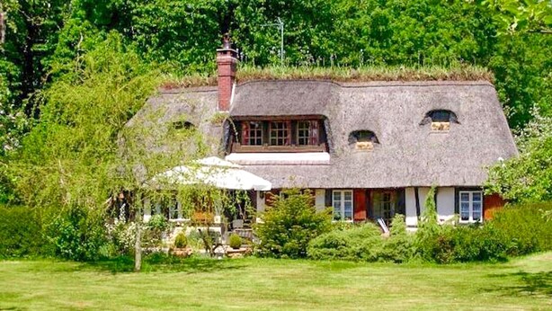 Un petit chalet au toit de chaume, doté d’une cheminée et entouré d’une végétation luxuriante.