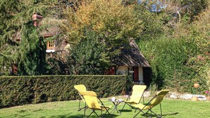 Un petit chalet au toit de chaume, avec un petit jardin, deux chaises pliantes jaunes et un arbuste au premier plan.