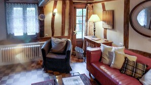 Un salon chaleureux avec un canapé rouge, un fauteuil sombre et un plancher en bois. Il y a deux fenêtres avec des rideaux, un radiateur et une lampe sur une table d’appoint.