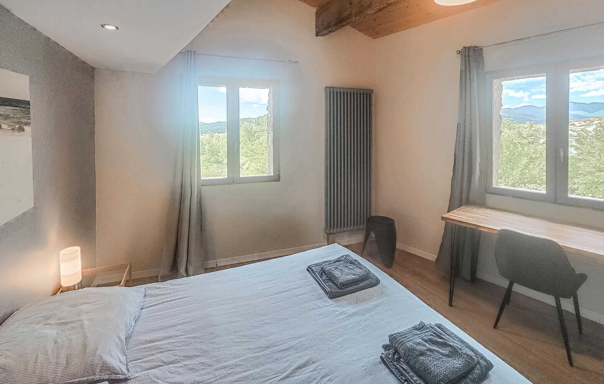 A bedroom with a bed, a desk, a chair, a radiator, and a window with a view.
