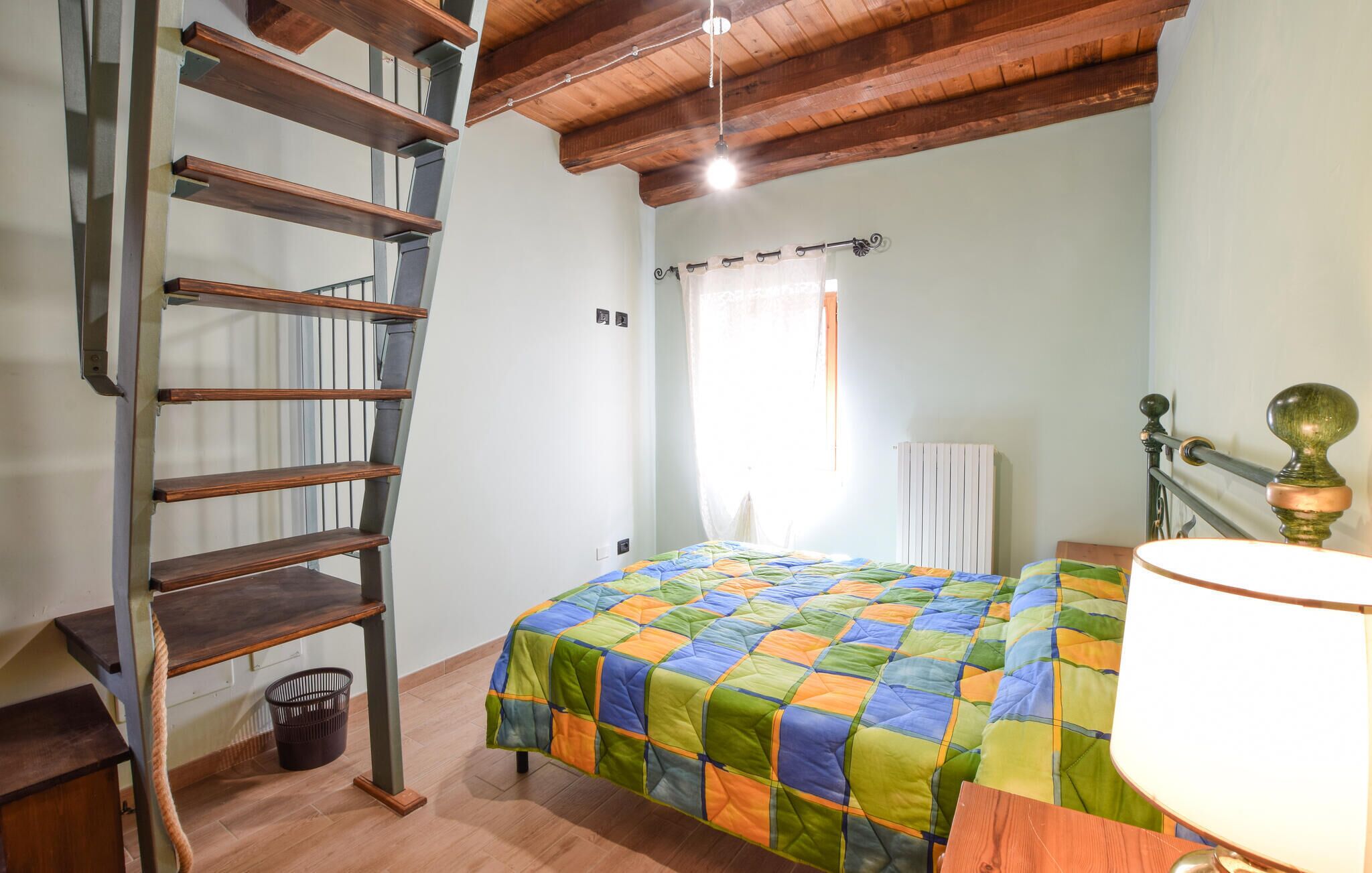A bedroom with a wooden ladder, a bed with a colorful quilt, a radiator, and a window with curtains.