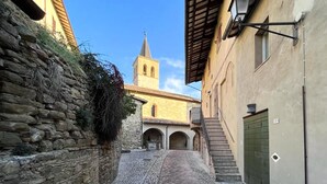 A narrow cobblestone street flanked by stone walls and buildings with arched doorways, leading to a church with a bell tower.