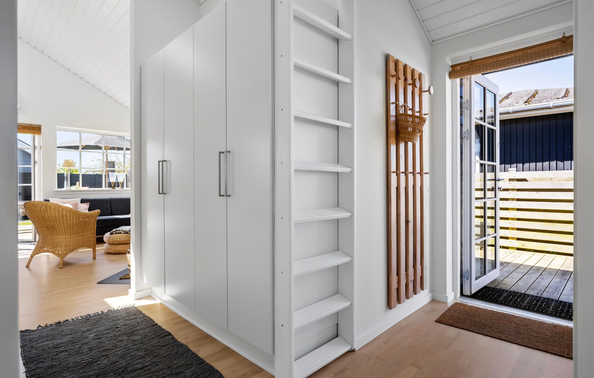 Un intérieur moderne avec une armoire blanche, un fauteuil en osier et une vue sur une terrasse en bois à l’extérieur.