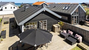 A modern house with a sloped roof, a wooden deck, and outdoor seating. - 2 bedroom cozy home in Otterup (Otterup)