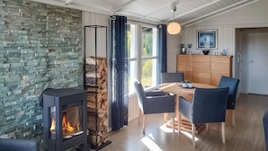 A cozy room with a stone wall, a wood-burning stove, a dining table with chairs, and a wooden cabinet with a framed picture. - Beautiful home in Trøgstad (Trøgstad)