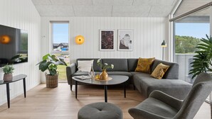 A modern living room with a grey sectional sofa, a round coffee table, and a flat-screen TV. There are two framed artworks on the wall, a floor lamp, and a large window offering a view of the outdoors.