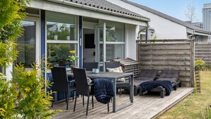 En overdækket terrasse med udendørs møbler, en træterrasse og udsigt til et hus med store vinduer.