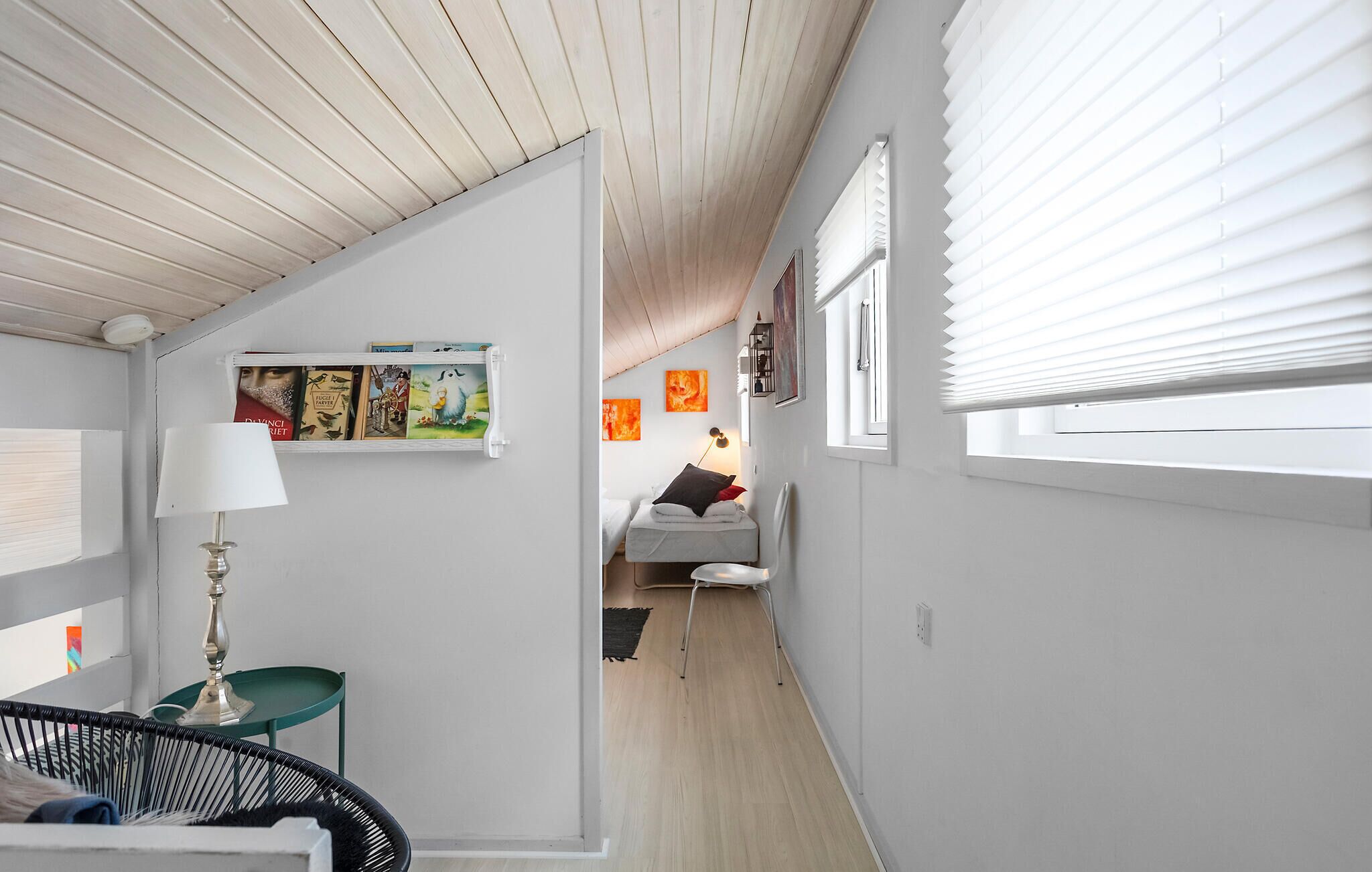 Un pasillo estrecho con una cama, una silla y una mesa con una lámpara. Hay cuadros en las paredes y una ventana con persianas.