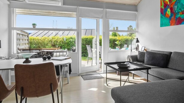 En moderne stue med en grå sofa, et spiseområde med bord og stole, og en stor skydedør der fører ud til en balkon med grønne planter.