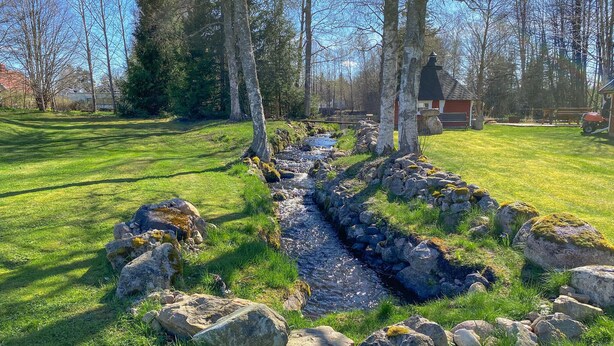 A rural landscape with a stone-lined stream, a wooden bench, and a small red building with a black roof. - Cozy home in Ormaryd with WiFi (Ormaryd)