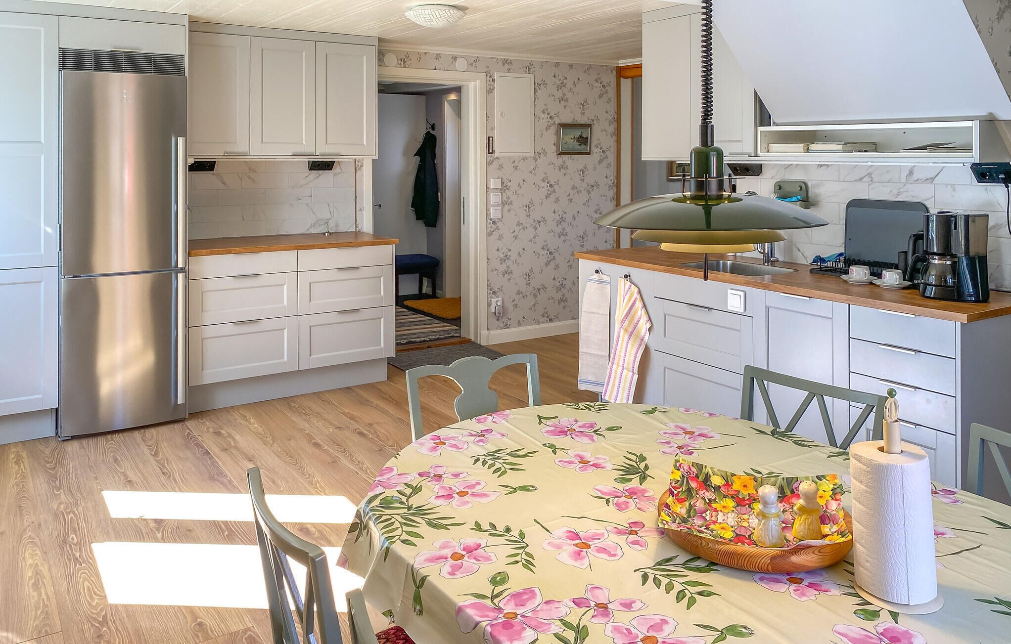 A compact kitchen with white cabinets, a stainless steel refrigerator, a wooden countertop, and a dining table with a floral tablecloth.