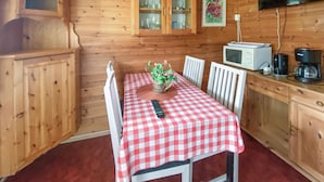 Puinen mökki, jonka sisätiloissa on ruokapöytä, joka on peitetty punavalkoisella ruutukuvioisella pöytäliinalla. Lisäksi siellä on valkoinen tuoli, pieni ruukkukasvi, kaukosäädin, valkoinen mikroaaltouuni ja kahvinkeitin.