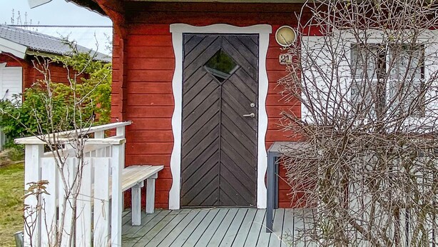 Une maison rouge avec une porte gris foncé, un porche blanc avec un banc et un petit arbre devant.