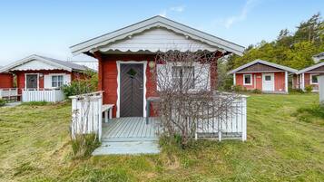 Une petite maison de plain-pied, à l’extérieur rouge et aux ornements blancs, dotée d’un porche avec un banc et d’un petit arbre devant.