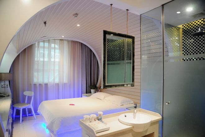 A modern hotel room with a curved ceiling, a large bed, a bedside table with a lamp, a chair, a large mirror, and a glass shower enclosure.