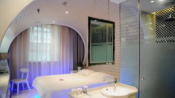 A modern hotel room with a curved ceiling, a large bed, a bedside table with a lamp, a chair, a large mirror, and a glass shower enclosure.