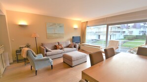 A modern living room with a sofa, ottoman, armchair, and dining table. A television is on a stand, and there is a window with a view of buildings and a garden. - Apartment in Seaside Bruges With Balcony (Knokke-Heist)
