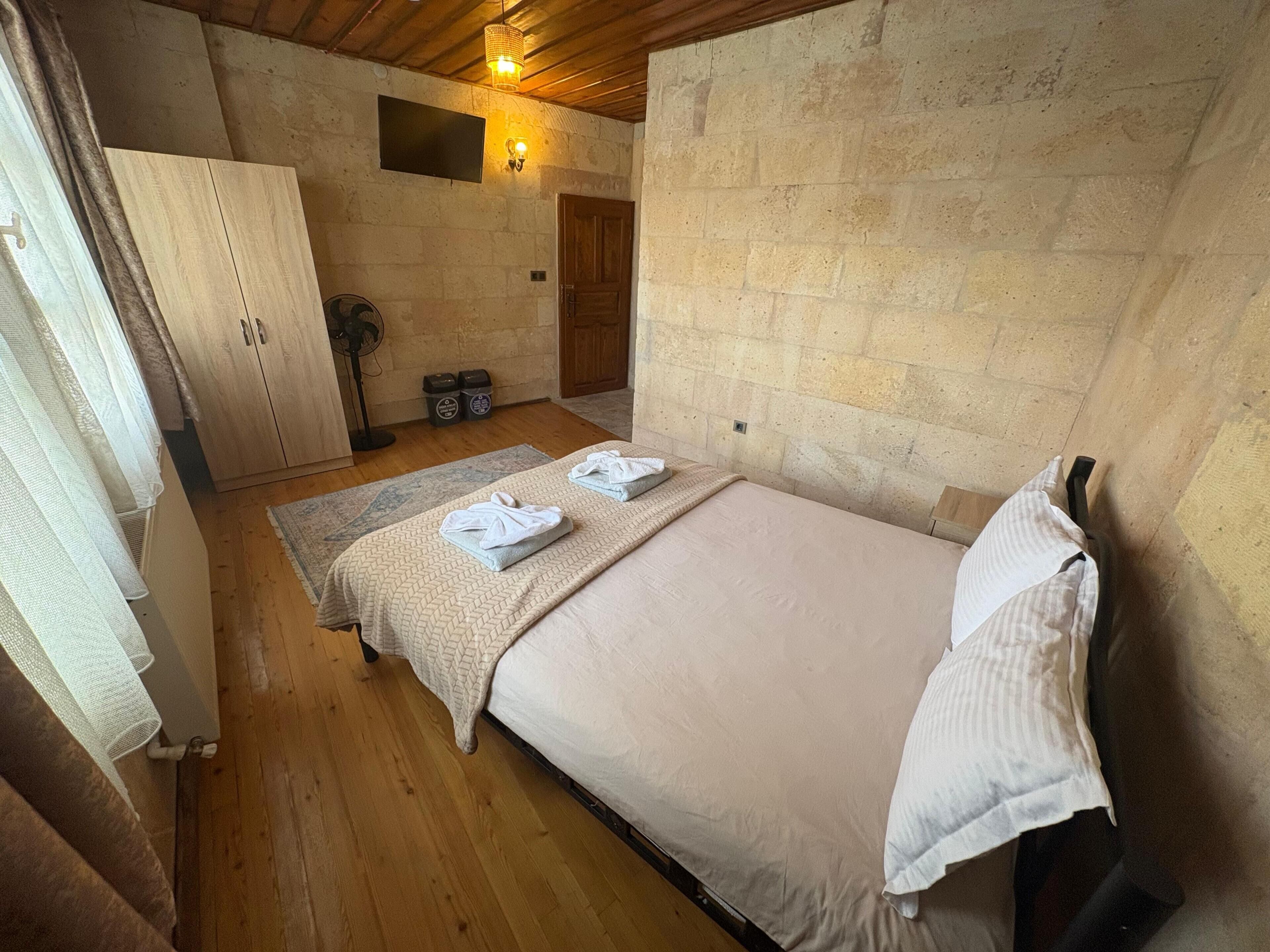 Una habitación con cama, un armario de madera, una mesita con toallas, un ventilador y una pared de piedra.