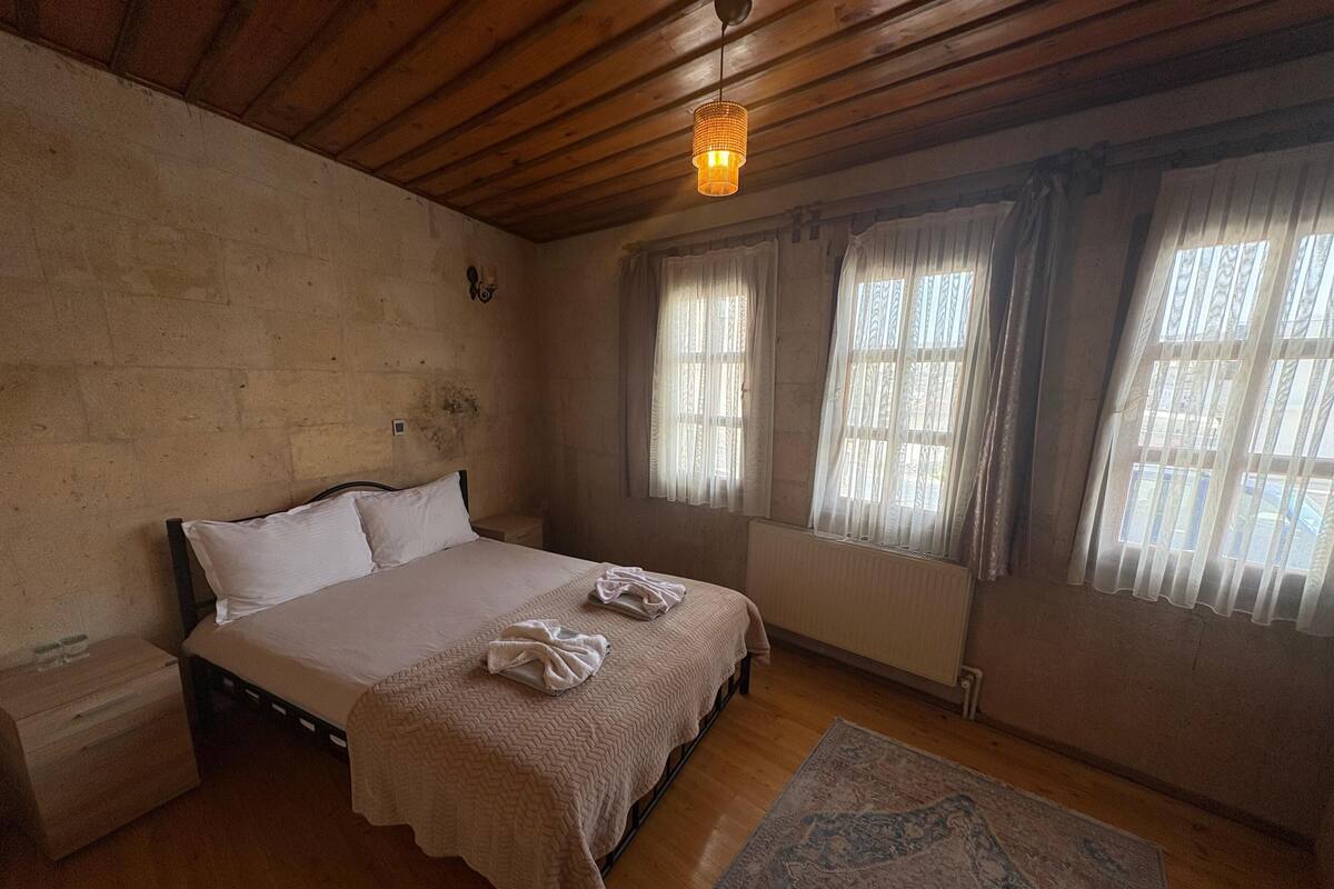 Un dormitorio con techo de madera, una cama con ropa de cama blanca, dos ventanas con cortinas translúcidas y piso de madera.