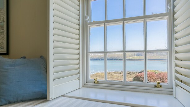 Una habitación con un ventanal grande que da a un lago, una almohada azul en el suelo y un cuadro en la pared.