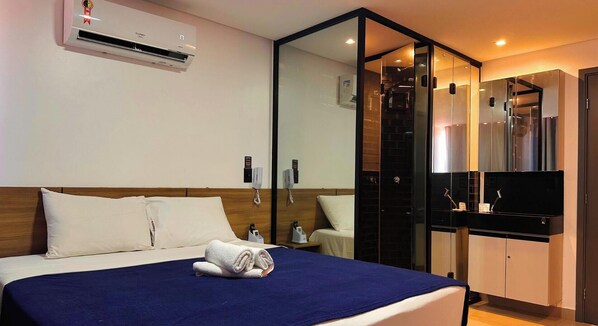 A hotel room with a bed, a blue bedspread, a white pillow, a white blanket, a towel, a wall-mounted air conditioner, a phone, a mirror, a black cabinet, and a television. - Get One (Joao Pessoa)